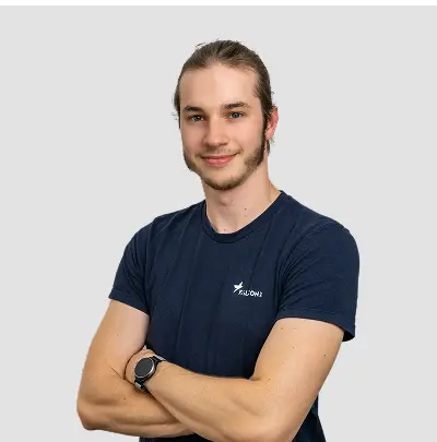 Junger Mann mit langen Haaren steht mit verschränkten Armen, trägt ein schwarzes T-Shirt mit Logo und eine Uhr, auf weißem Hintergrund.