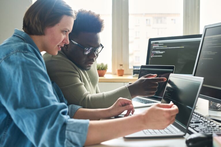 Zwei Personen arbeiten gemeinsam an Computerprogrammen, während sie auf mehrere Bildschirme mit Code schauen. Der Raum ist hell erleuchtet und es sind verschiedene Laptops und Computer sichtbar.